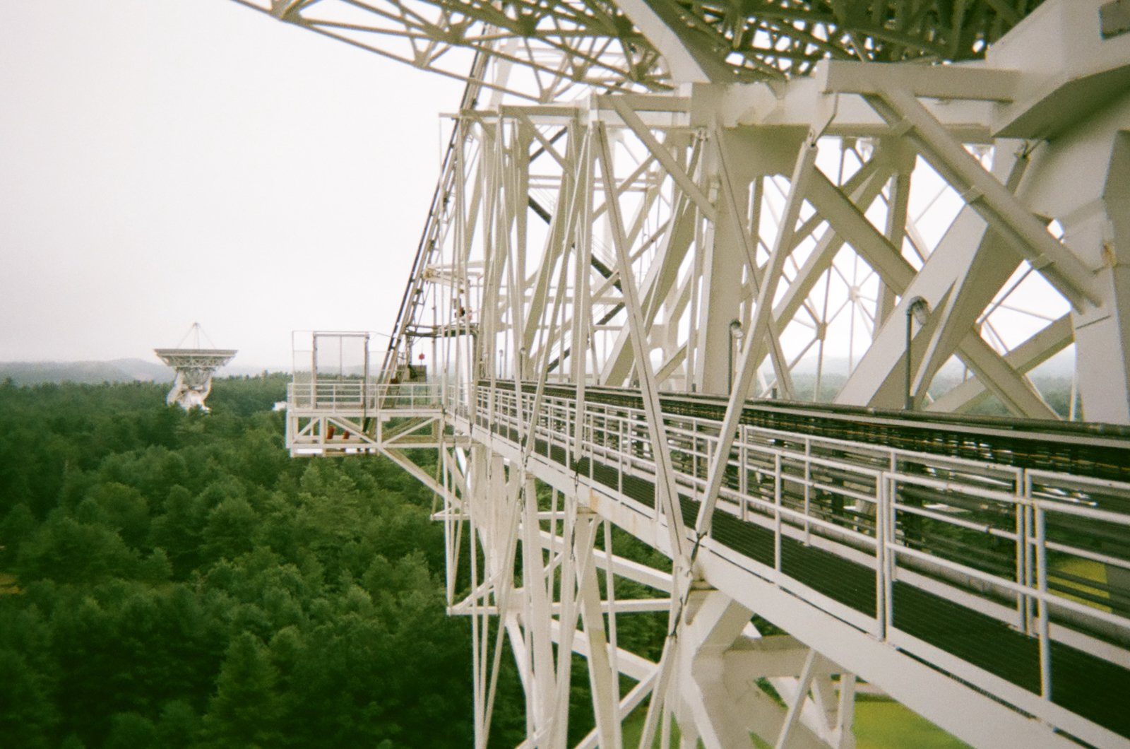 View from high on the structure of the Robert C. Byrd Green Bank Telescope, looking along a steel walkway with safety railings. The white lattice of the dish frame fills the upper frame. In the distance, a second smaller dish stands above a dense forest canopy under flat, diffuse light.