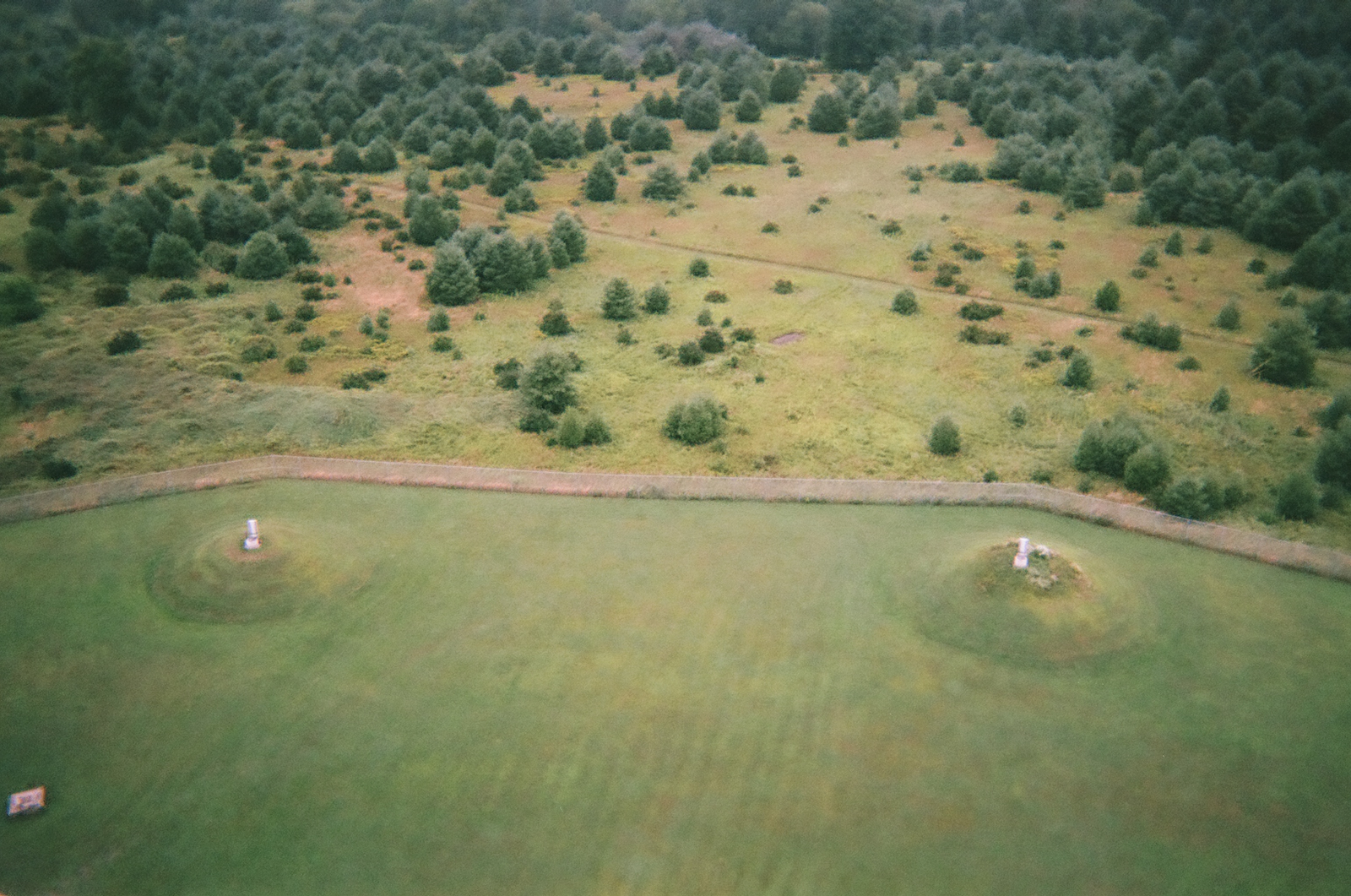 Photograph taken at roughly a 45-degree angle from the Robert C. Byrd Green Bank Telescope, looking down and out across the observatory grounds. Two circular mounds of grass, each topped with a small white laser alignment post, sit symmetrically in a trimmed field divided by a gravel path. Scattered conifers and scrubland stretch back toward forested hills.
