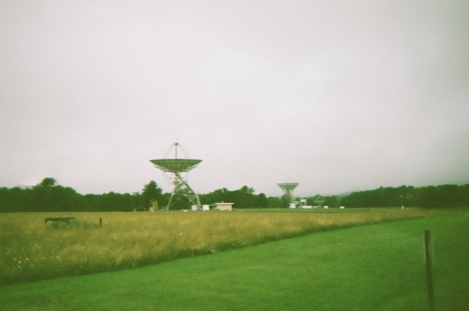 Disposable camera photograph of two radio telescopes rising above a flat field at the Green Bank Observatory, West Virginia, under a heavy overcast sky. Bright mown grass in the foreground gives way to tall wild grass, with a treeline behind the telescopes. The larger dish faces upward, pale against grey.